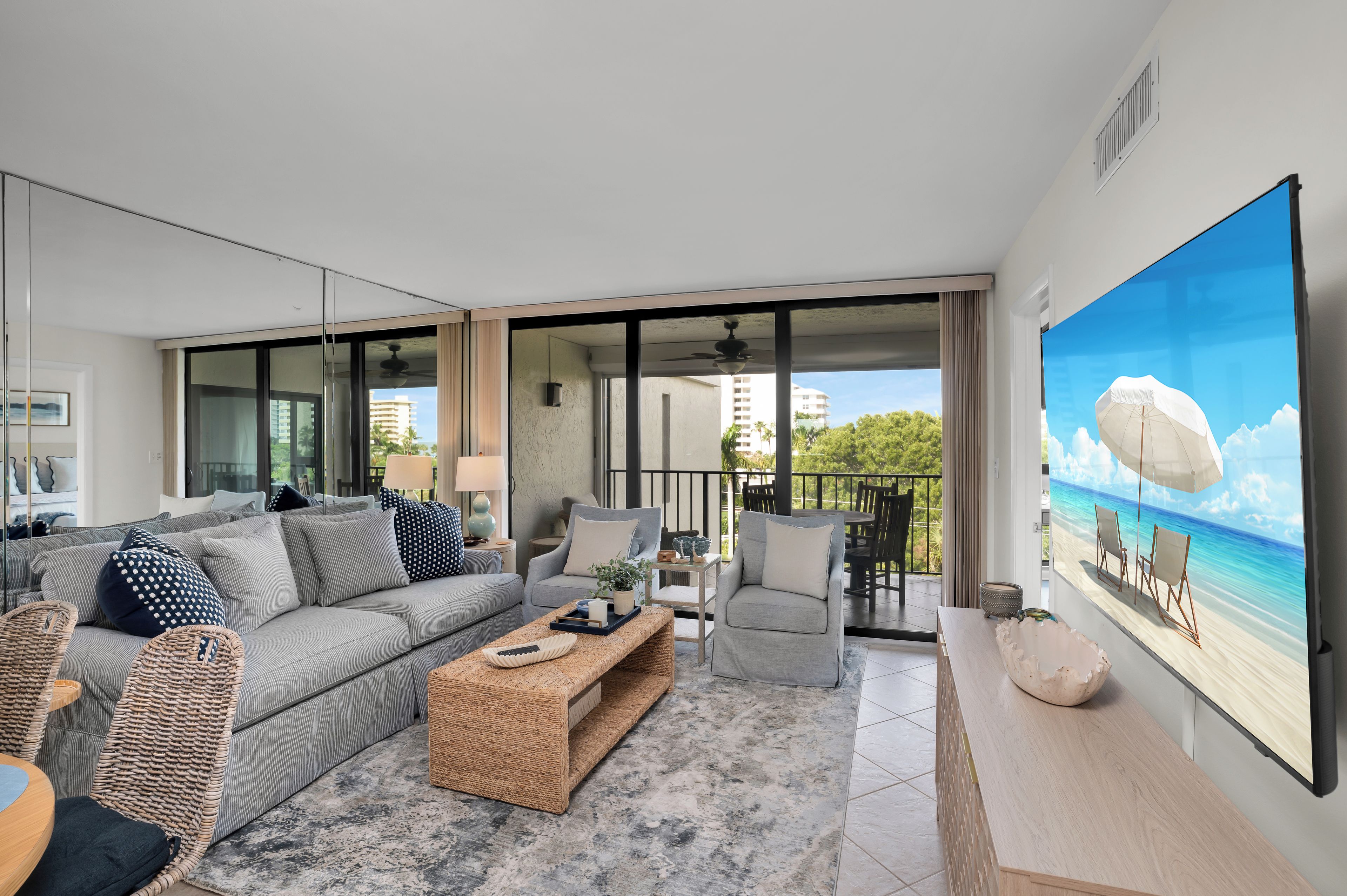 A modern living room with a large flat-screen TV, a grey sofa, a wicker coffee table, and a balcony with a view of the ocean.
