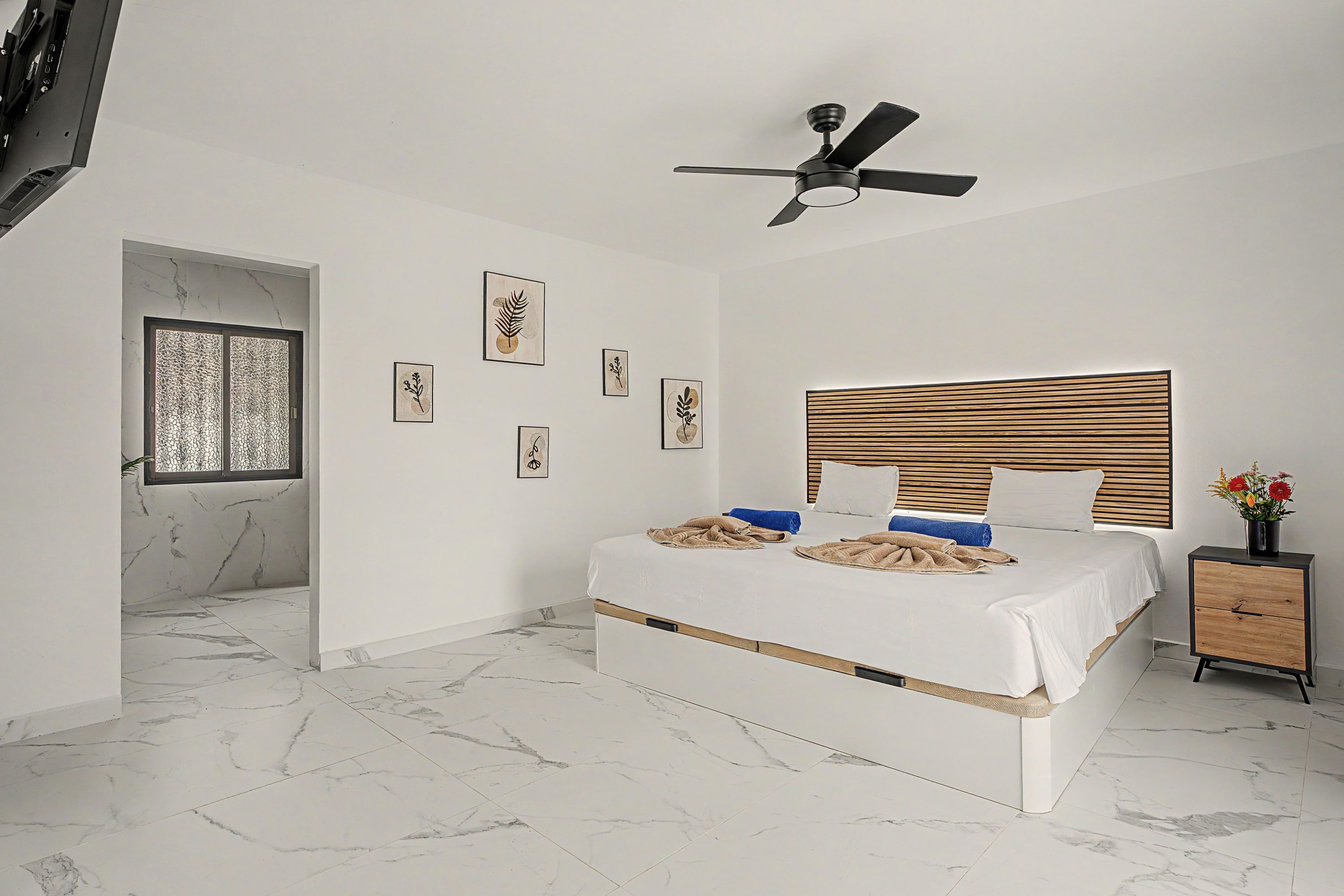 Un dormitorio con cama, ventilador de techo, cabecera de madera, mesita de noche y ventana con celosía decorativa.