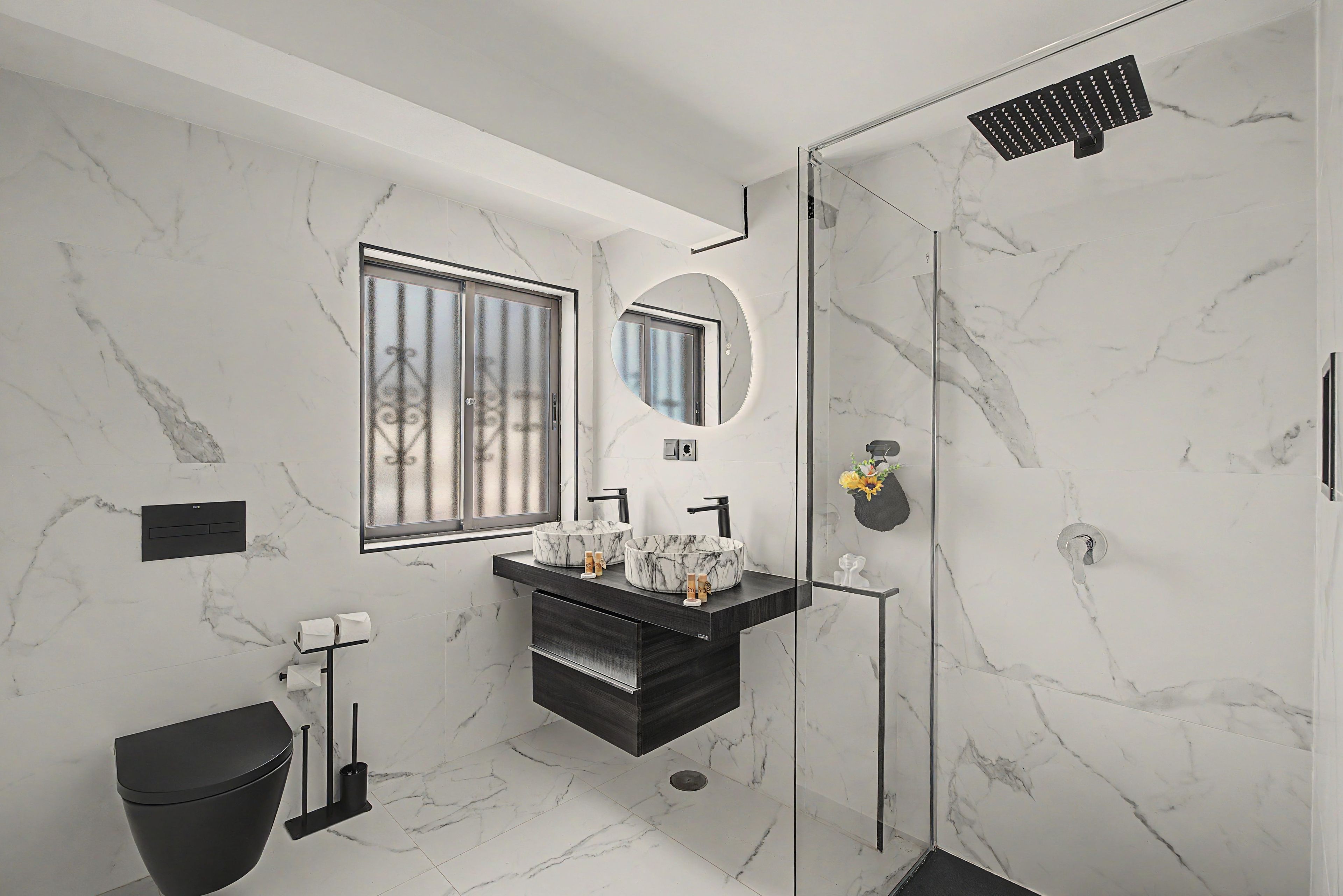 Un baño moderno con piso de mármol, tocador negro, cabina de ducha de vidrio y ventana con vistas.