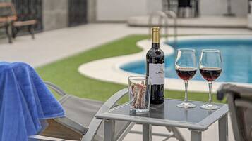 Escena junto a la piscina con una botella de vino, dos copas y un vaso con una bebida.