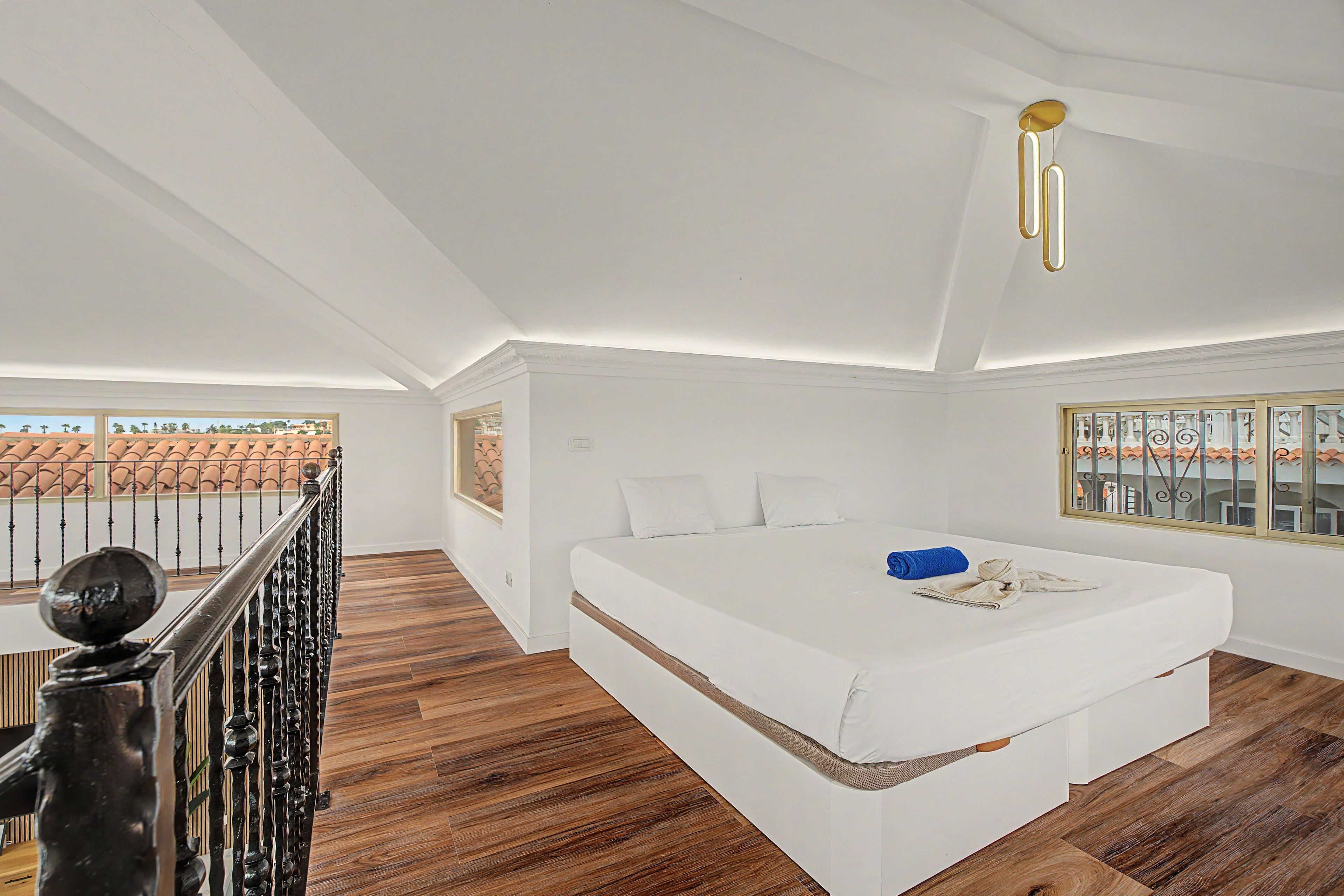 Un dormitorio con una cama grande, suelo de madera, un balcón con barandilla y vistas a edificios.