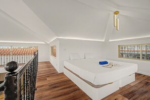 Un dormitori amb un llit gran, terres de fusta, un balcó amb barana i vista a edificis.