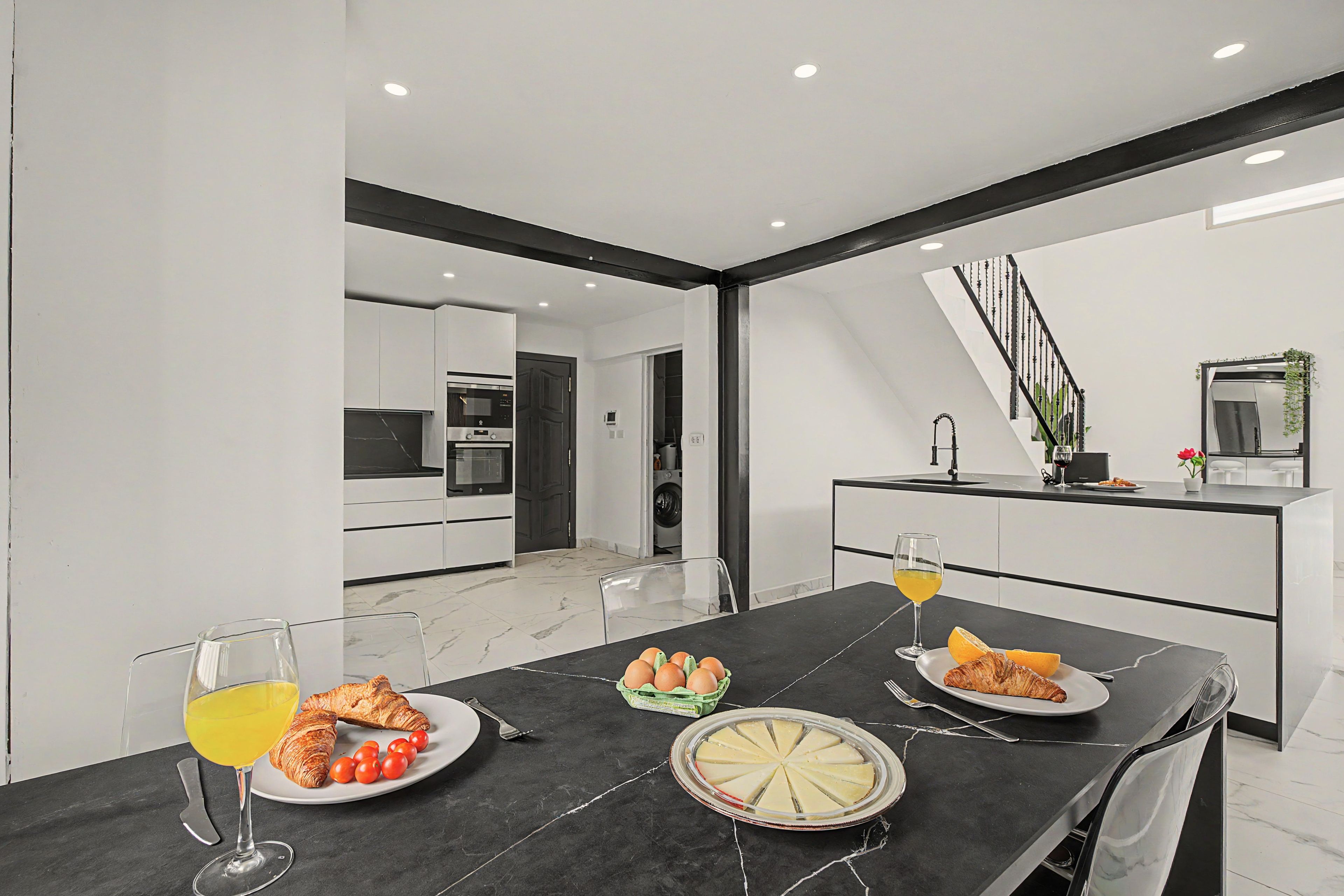 Un comedor moderno con una mesa de mármol negro, una cocina al fondo y una escalera que sube a otro nivel.