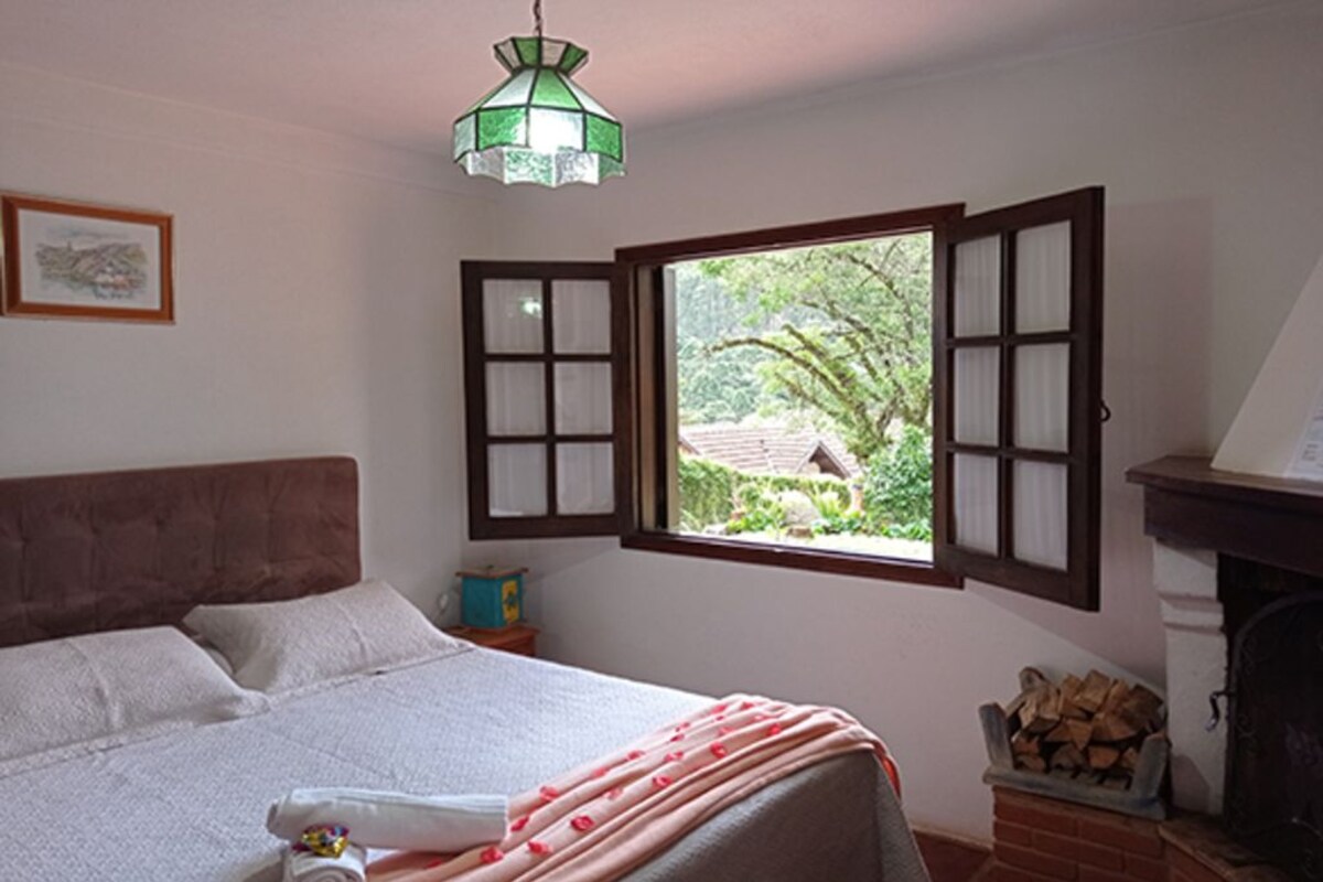 Une chambre à coucher avec un lit, une fenêtre dont les volets sont ouverts, une cheminée et un luminaire suspendu.