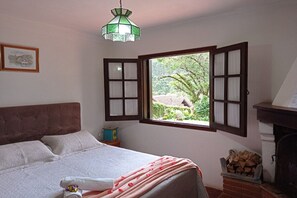 Une chambre à coucher avec un lit, une fenêtre dont les volets sont ouverts, une cheminée et un luminaire suspendu.