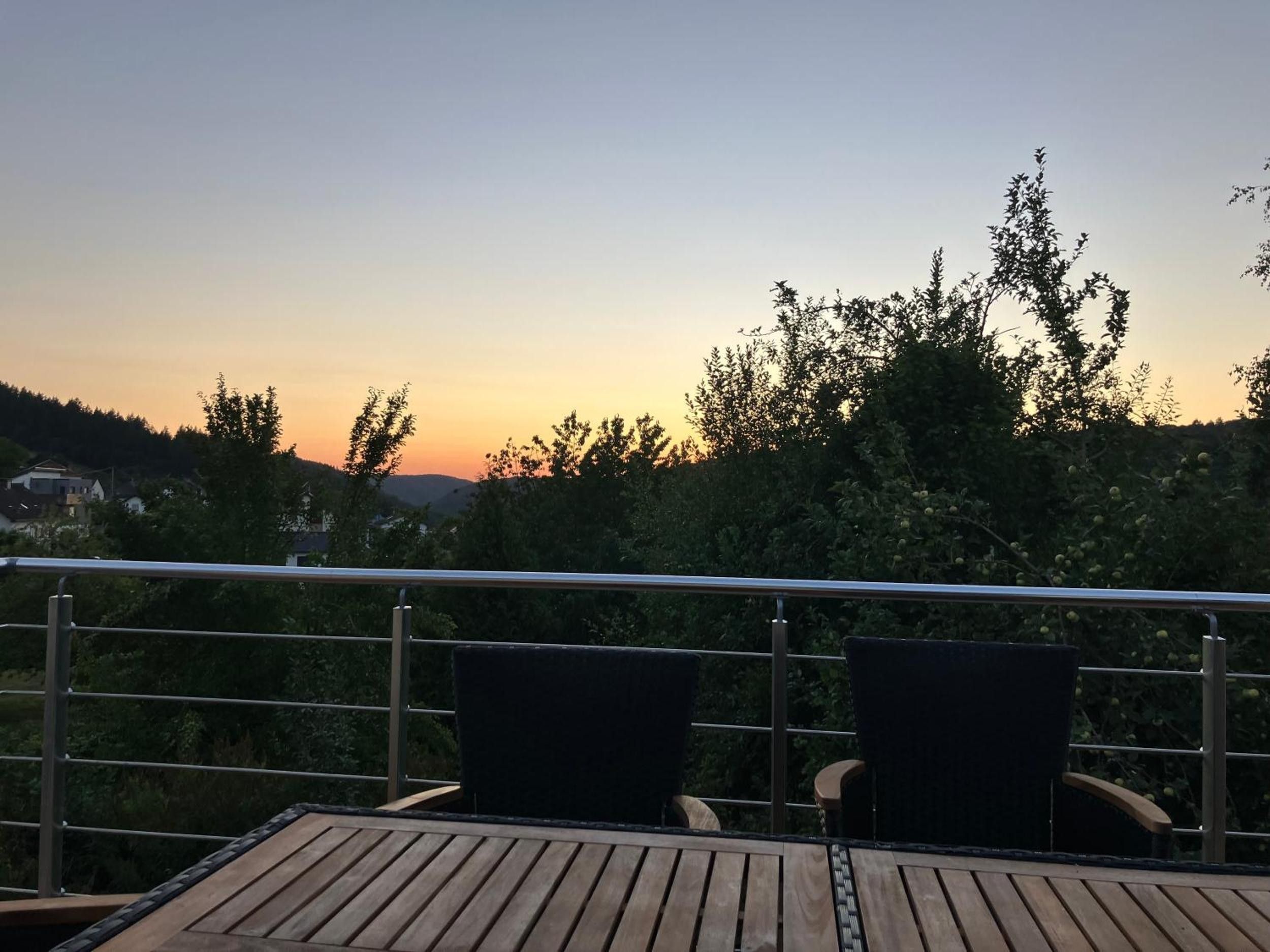 Eine Holzterrasse mit zwei Stühlen und einem Geländer, mit Blick auf einen Sonnenuntergang, Bäume und Hügel im Hintergrund.
