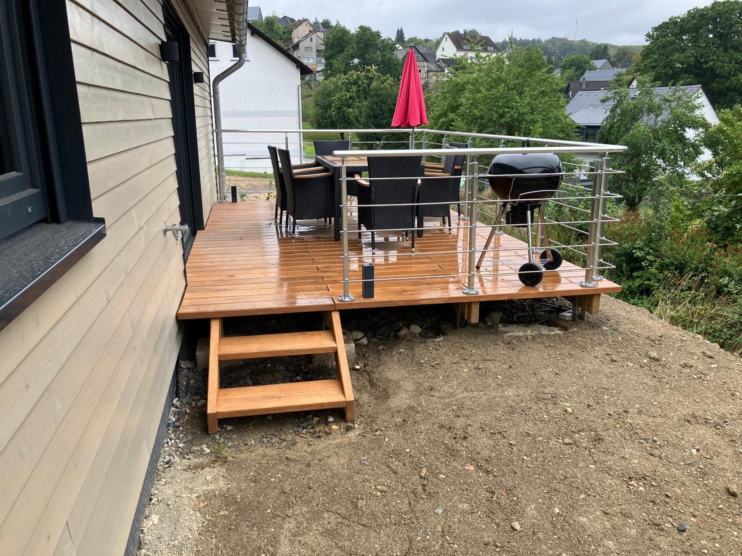 Eine Holzterrasse mit Gartenmöbeln, einem Grill und einem roten Sonnenschirm.