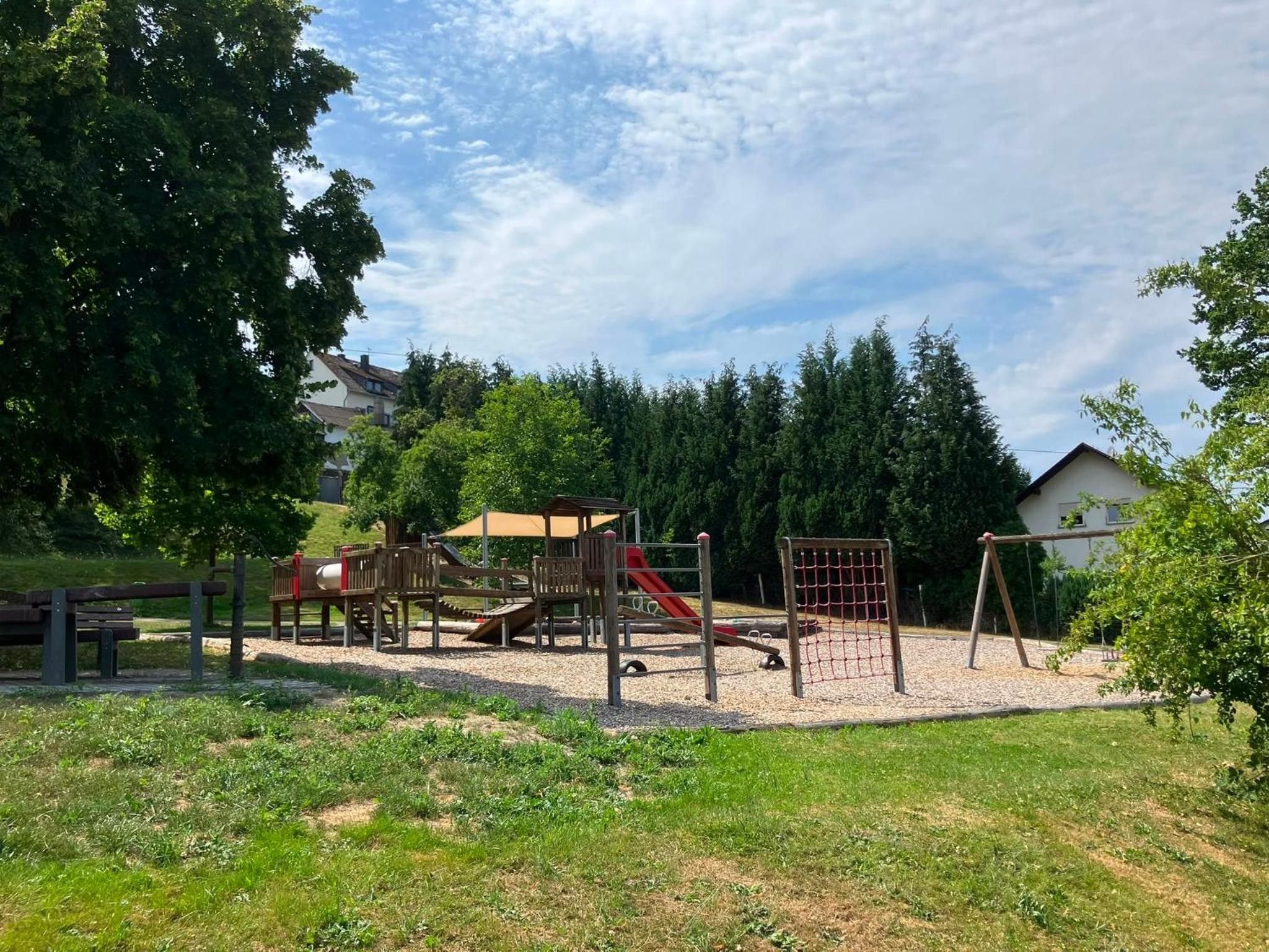Un parque infantil con diversas estructuras de juego, incluyendo un tobogán y juegos de escalada, rodeado de árboles y césped.