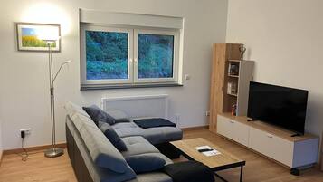 Ein modernes Wohnzimmer mit einer Couch, einem Couchtisch, einem Fernseher, einem Bücherregal und einem Fenster mit Blick auf Grünflächen.