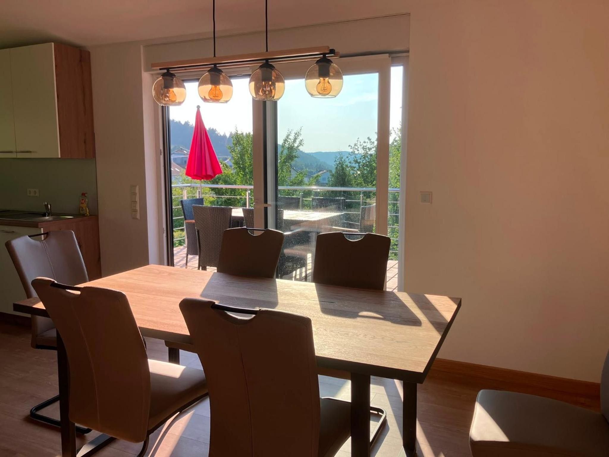 A modern dining area with a wooden table and chairs, a kitchen in the background, and a view of the outdoors through large windows.