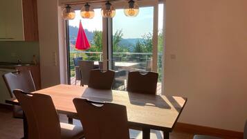 A modern dining area with a wooden table and chairs, a kitchen in the background, and a view of the outdoors through large windows.