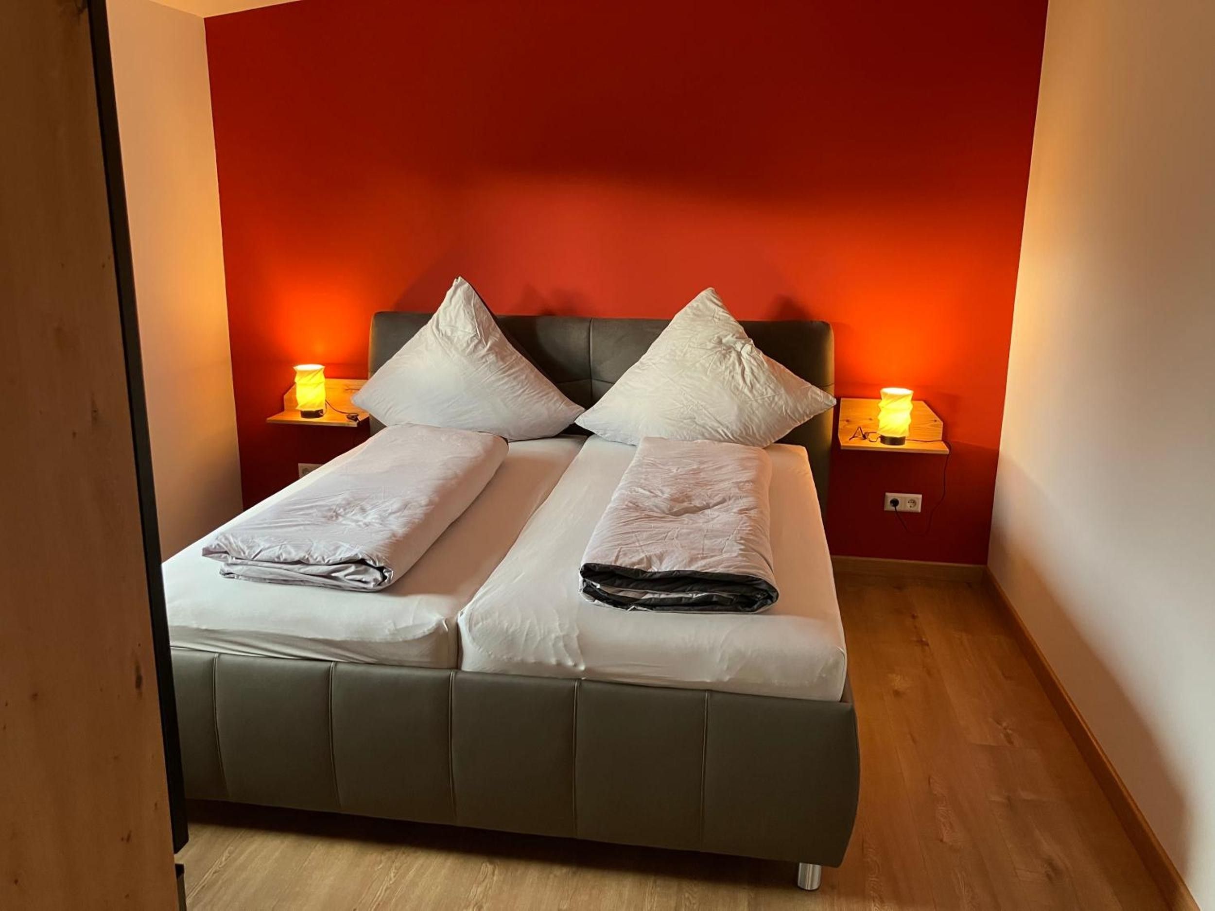 Ein Bett mit zwei Kissen und einer Decke, ein Nachttisch mit Lampe und eine rote Wand.