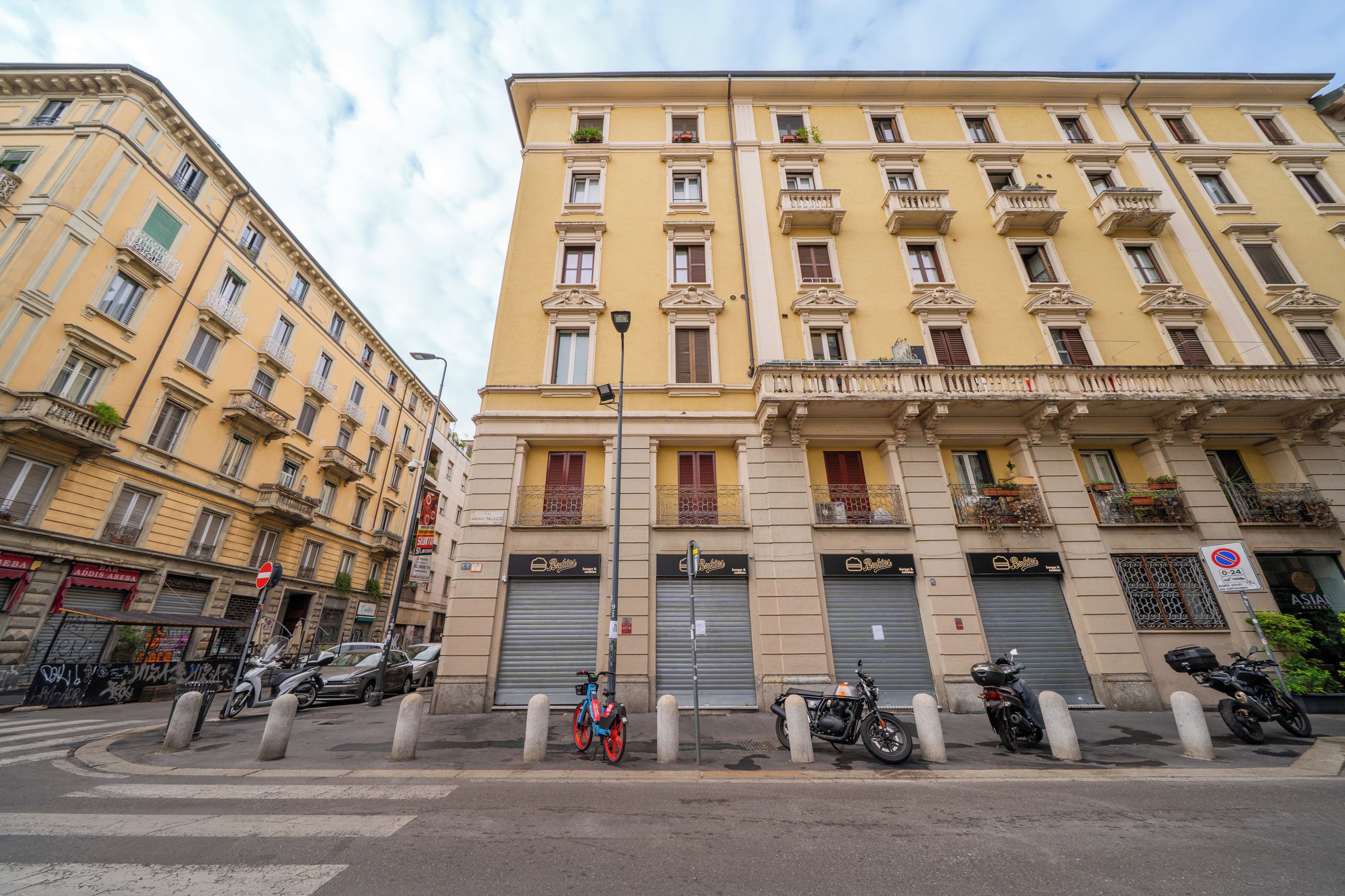 Un edificio a più piani con persiane chiuse, una strada delimitata da dissuasori e motociclette parcheggiate.