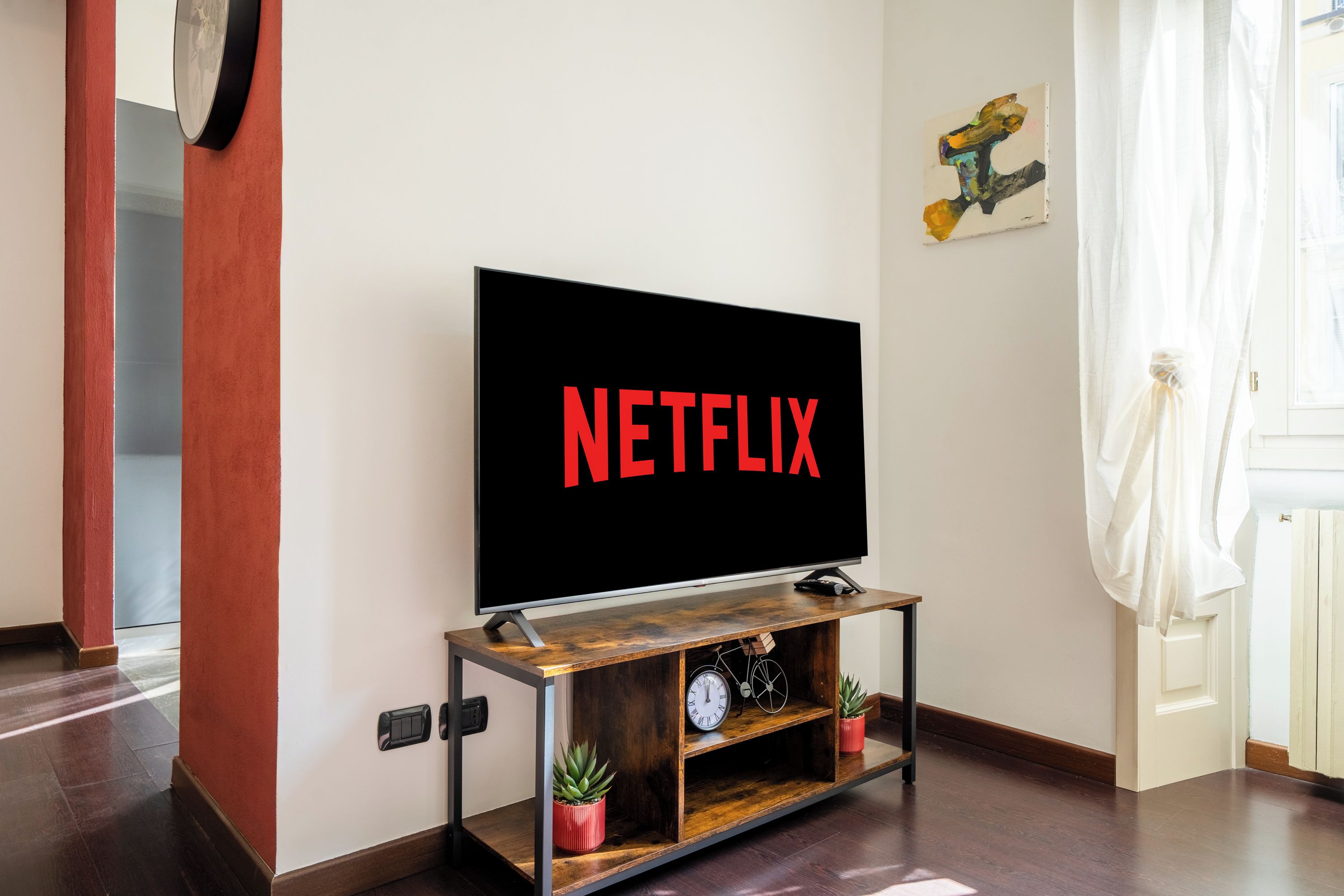 Una televisione su un supporto in legno con ripiano sottostante dove si trovano un orologio e una pianta.