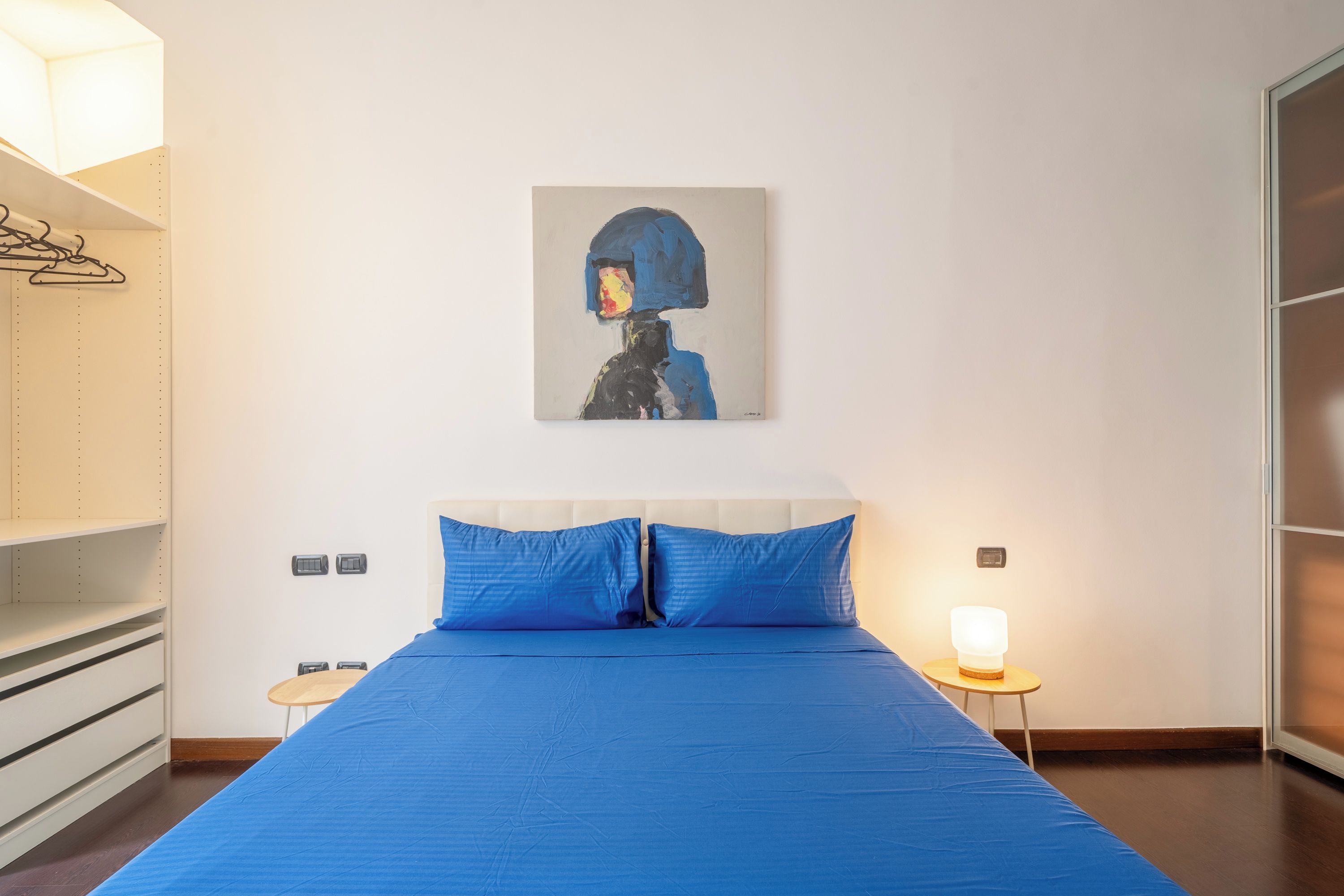 Una camera d'albergo con un letto, un'opera d'arte appesa al muro, un comodino con una lampada e un armadio.