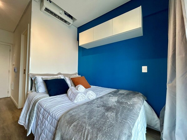 A hotel room with a bed, pillows, a blanket, an air conditioning unit, and a blue accent wall. - SETIN 2302 (São Paulo)