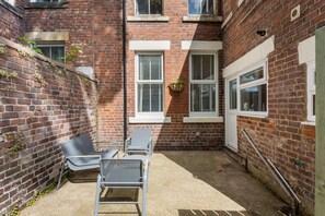 Ein Backsteinhaus mit kleiner Terrasse, zwei Fenstern mit Fensterläden und einer hängenden Pflanze.