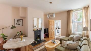 A cozy living room with a fireplace, a sofa, a dining table, and a kitchen visible through an open door.