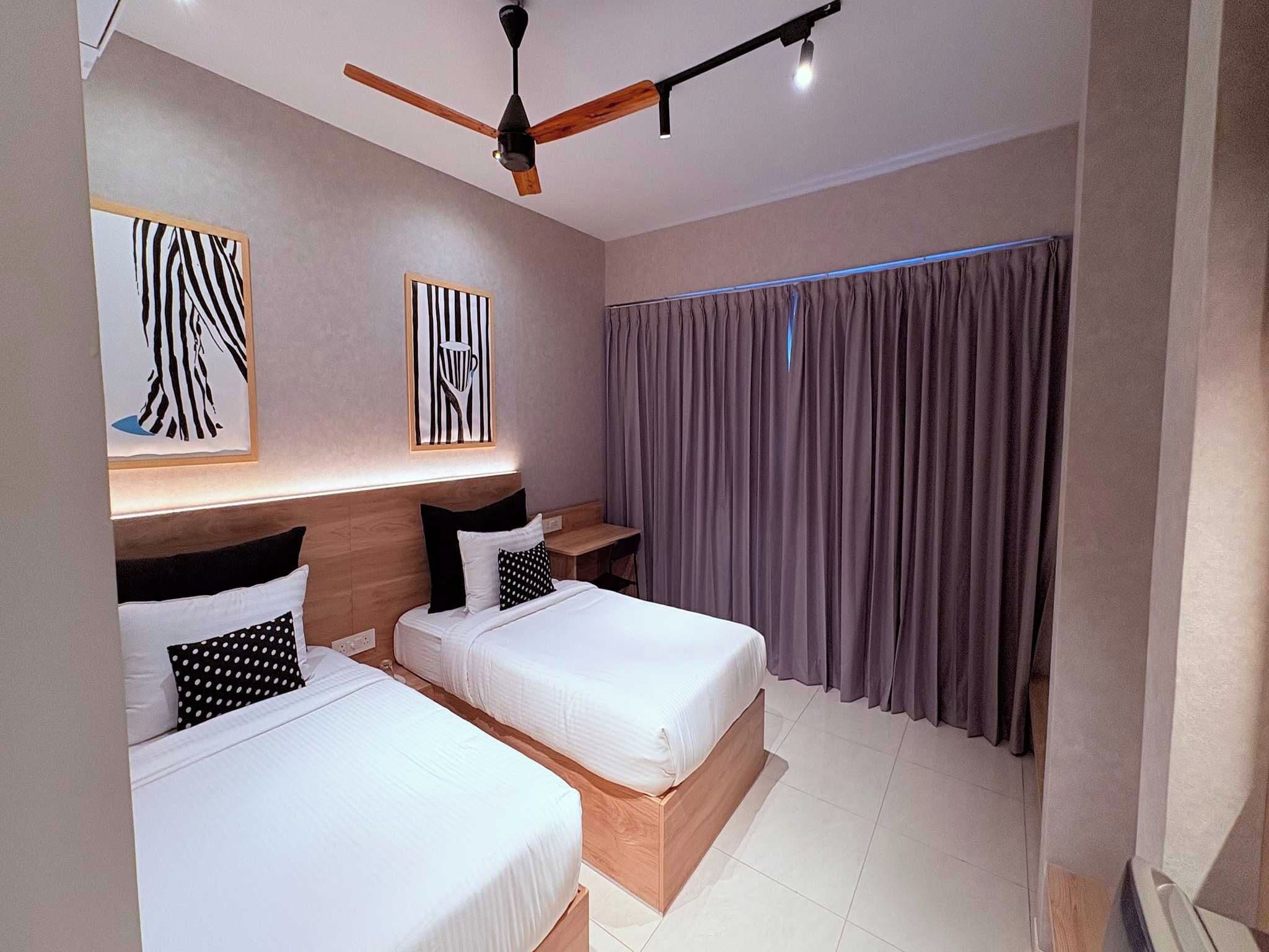 A hotel room with two beds, a ceiling fan, artwork on the walls, and a window with curtains.