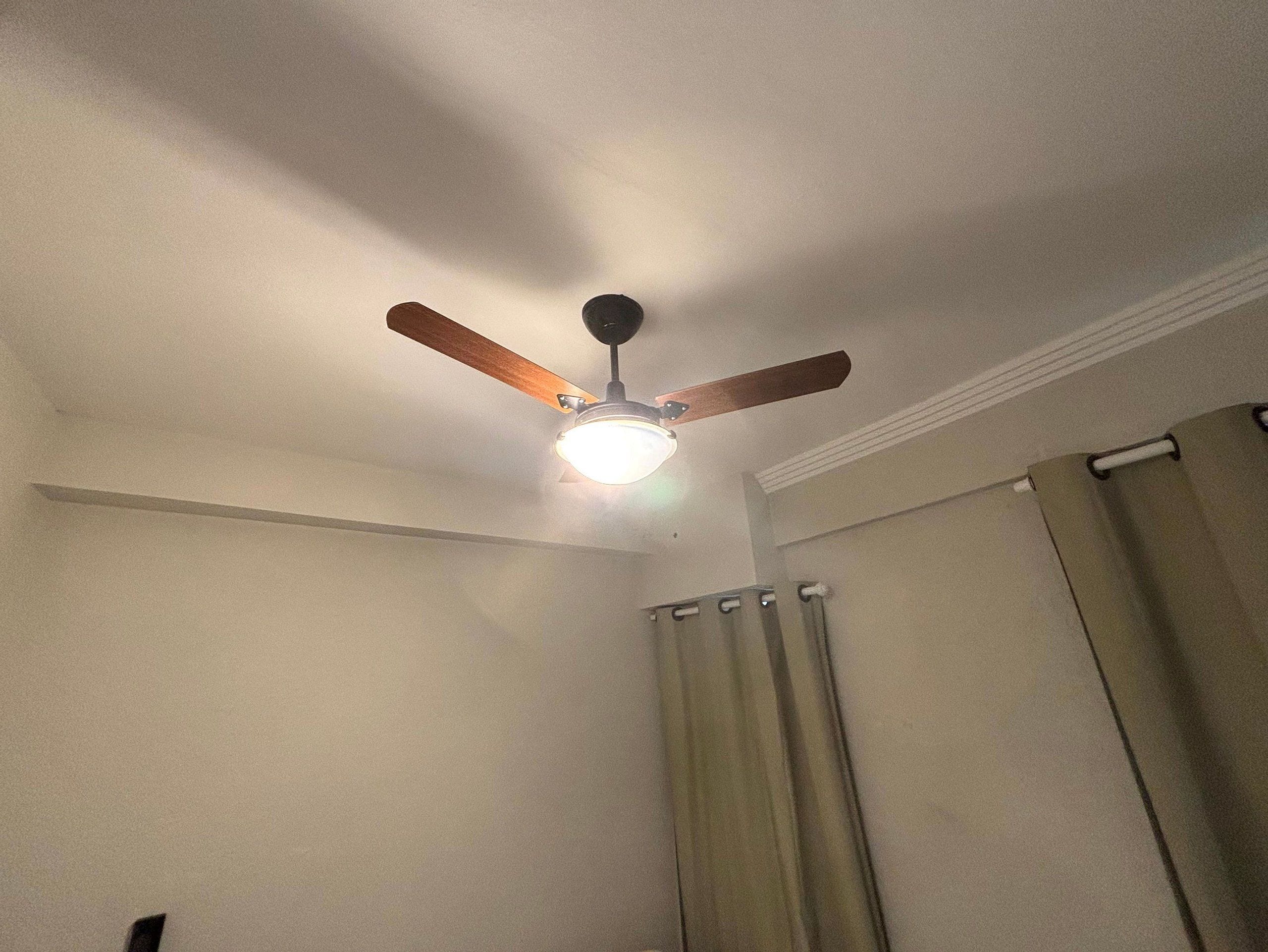 Un ventilador de techo con lámpara en una habitación con paredes lisas y una cortina.