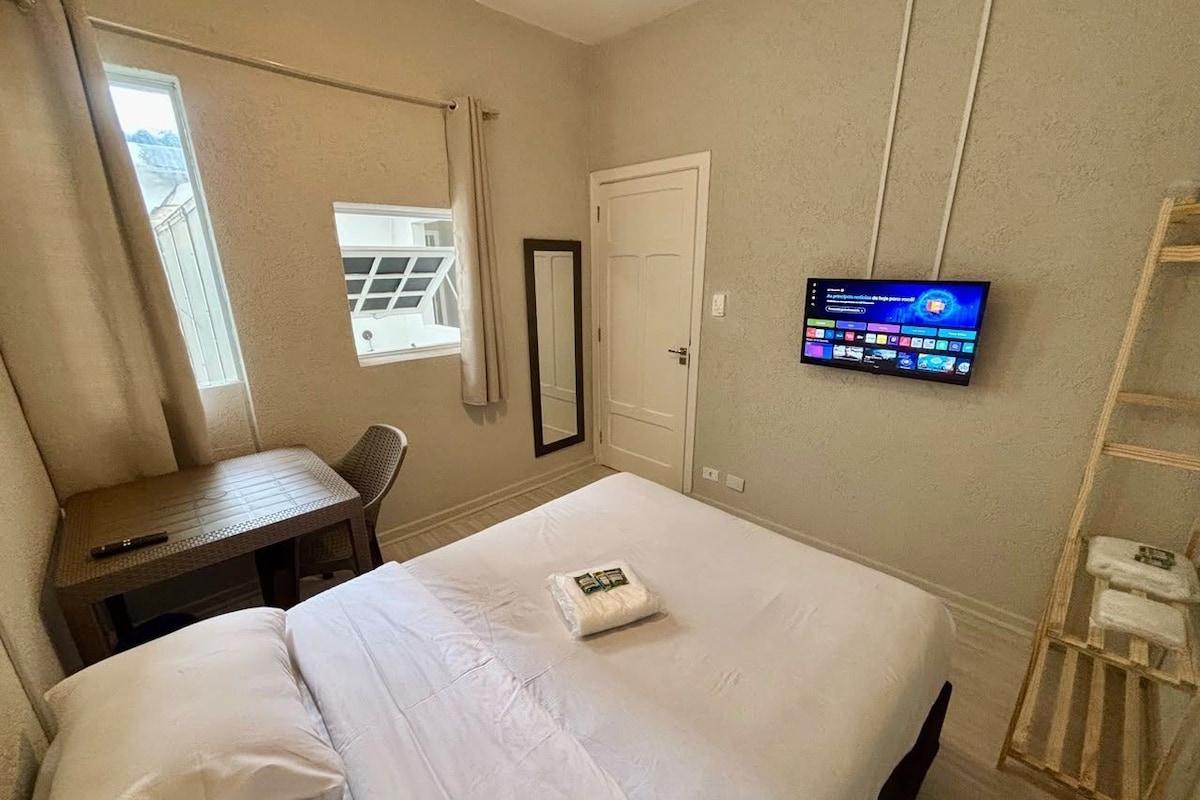 Quarto de hotel com cama, escrivaninha, televisão e janela.