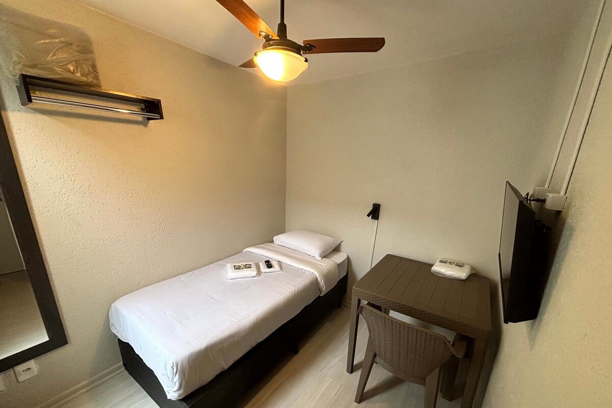 Quarto pequeno com cama de solteiro, ventilador de teto, mesa de cabeceira e uma cadeira.