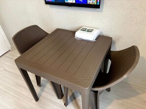 Uma mesa pequena e quadrada com tampo trançado e duas cadeiras de vime. Um travesseiro branco está sobre a mesa.