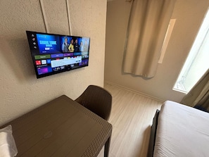 Quarto de hotel com televisão de tela plana na parede exibindo um canal esportivo, uma mesinha, uma cadeira e uma cama.