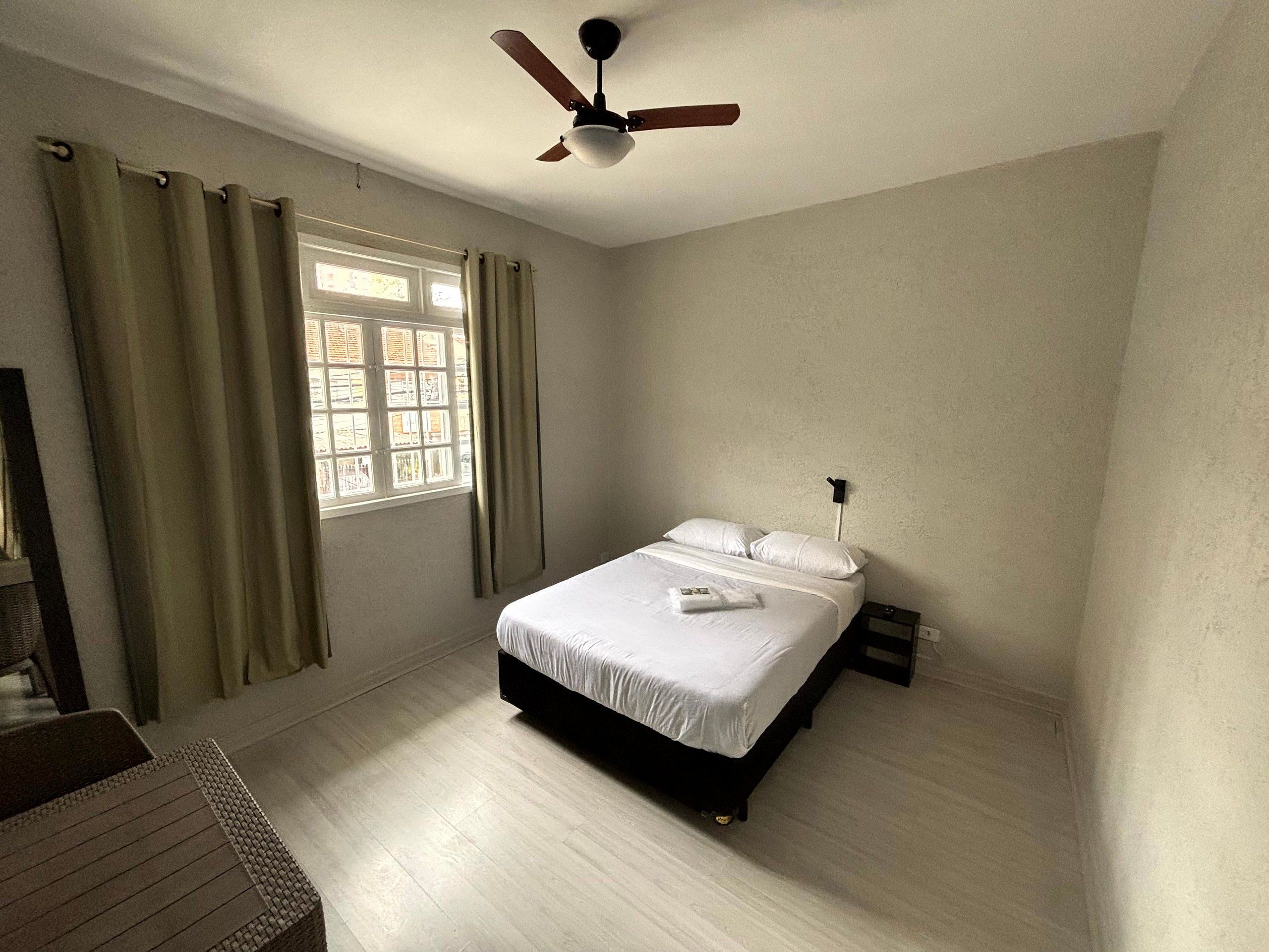 Un dormitorio con cama, ventilador de techo, ventana con cortinas y mesita de noche.