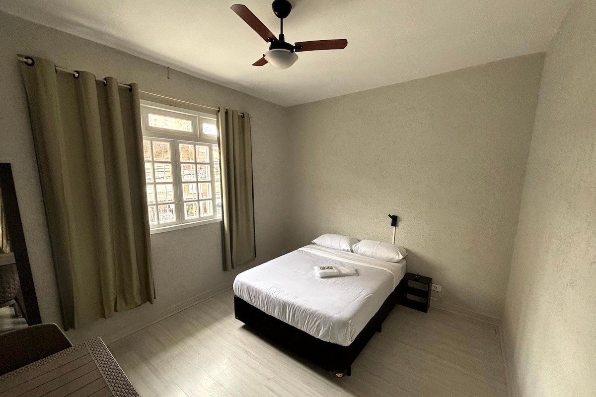 Quarto com cama, ventilador de teto, janela com cortinas e mesa de cabeceira.