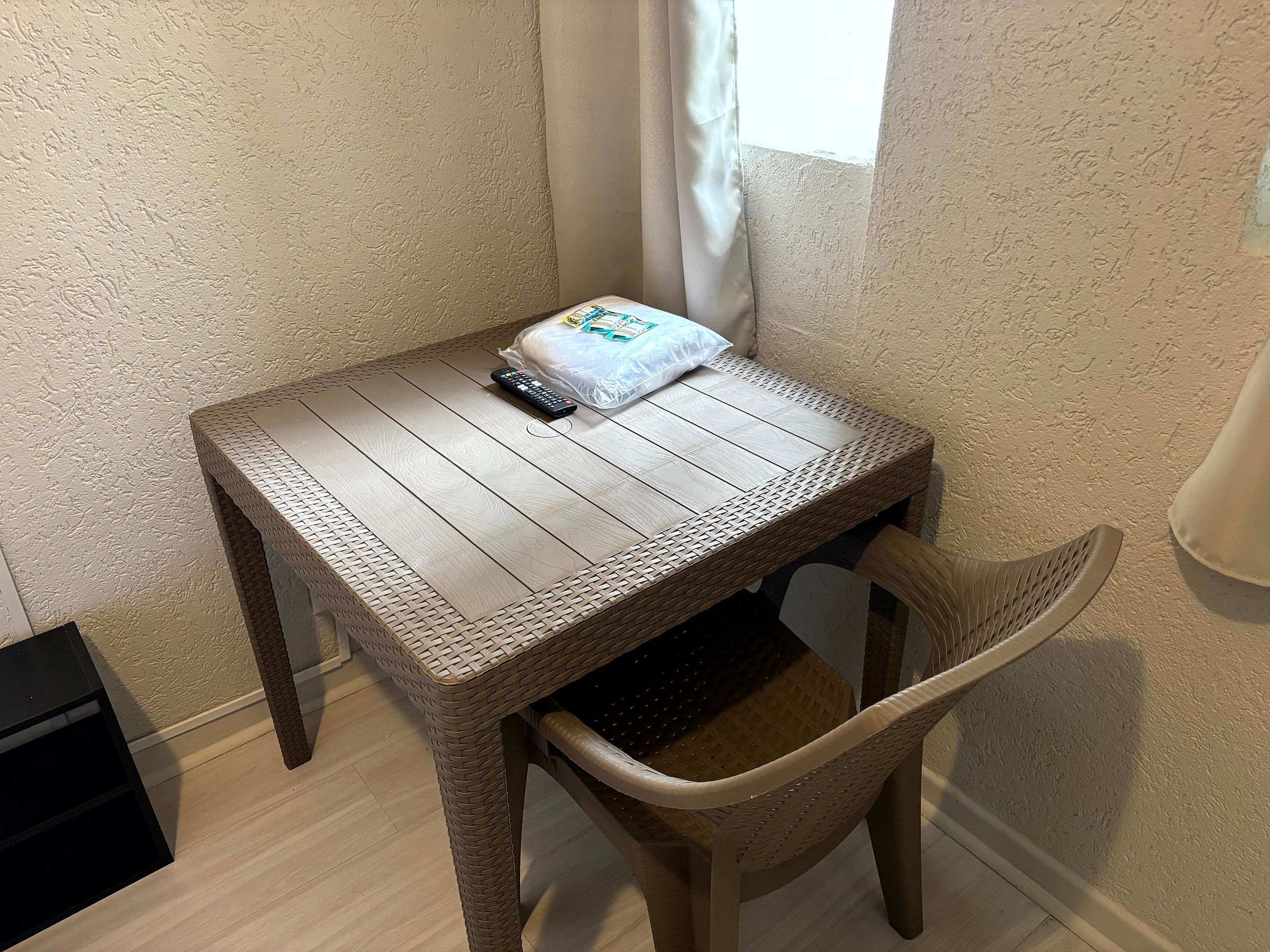 Una pequeña mesa de madera con un control remoto y un almohadón, una silla de mimbre y una ventana con cortinas.