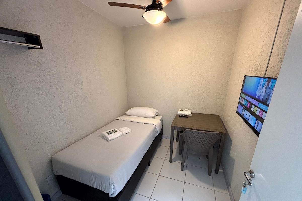 Quarto pequeno, com uma cama de solteiro, ventilador de teto, mesa e televisão fixada na parede.