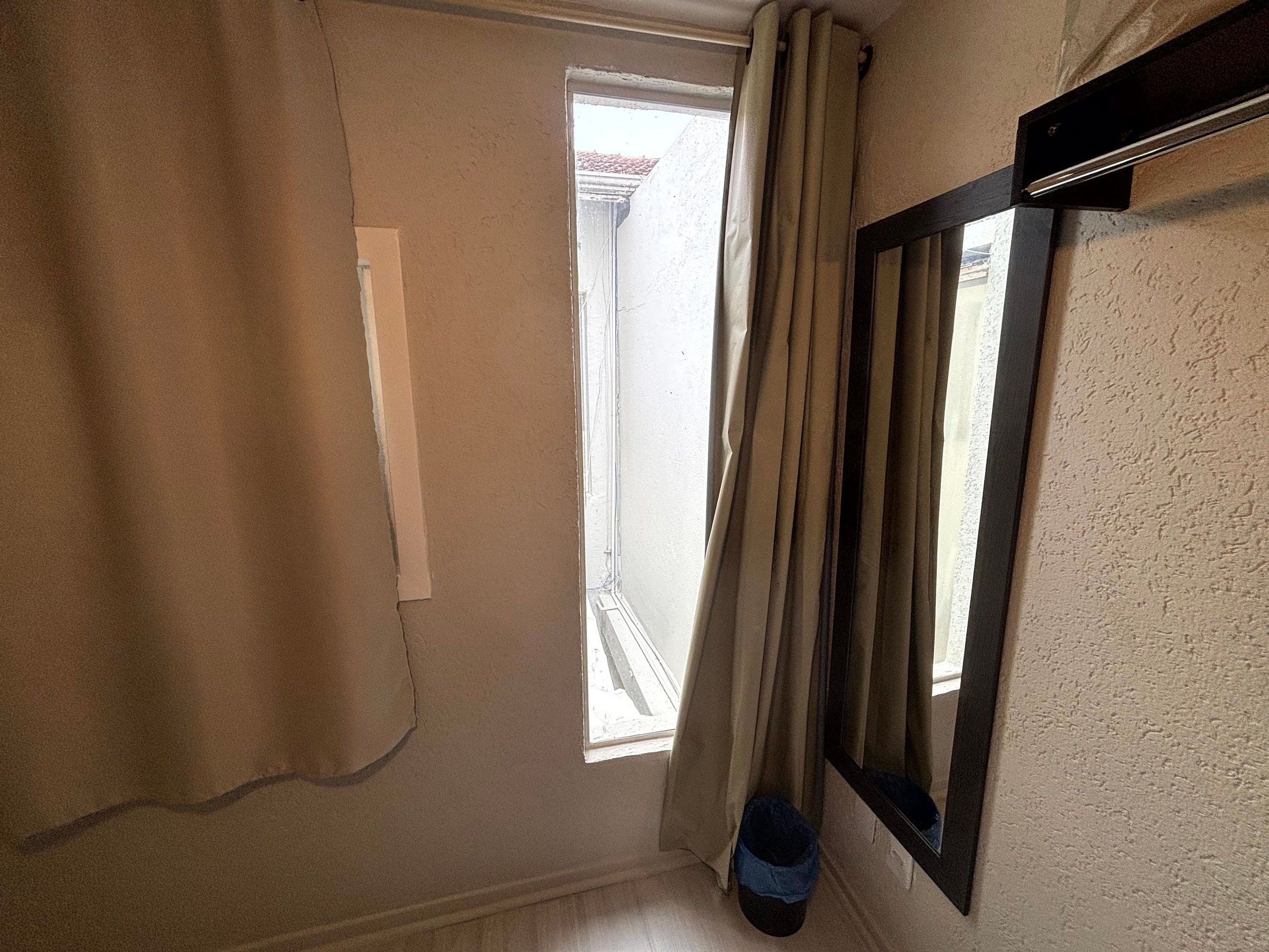 Una habitación con ventana, cortina, espejo y un par de zapatos en el suelo.