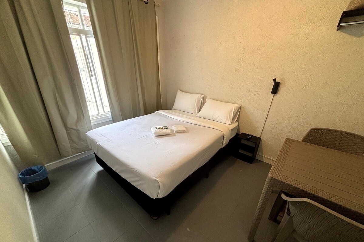 Cama bem arrumada com lençóis brancos e uma pequena mesa de cabeceira com abajur.