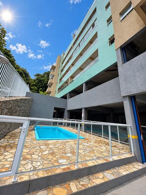 Un edificio de varios pisos con zona de piscina, suelo de piedra y cielo despejado.