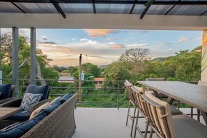 A covered outdoor seating area with a table and chairs, overlooking a lush landscape with trees and buildings. - New Listing • Dos Mangos • 3BR Walk to Beach • Playas del Coco (Playas del Coco)