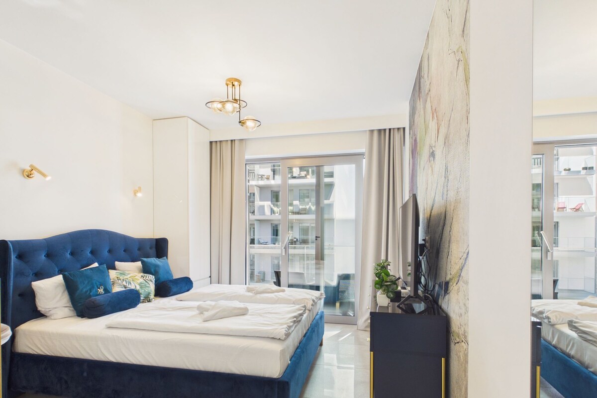 A modern bedroom with a large bed, a flat-screen TV, and a view of the building's exterior.