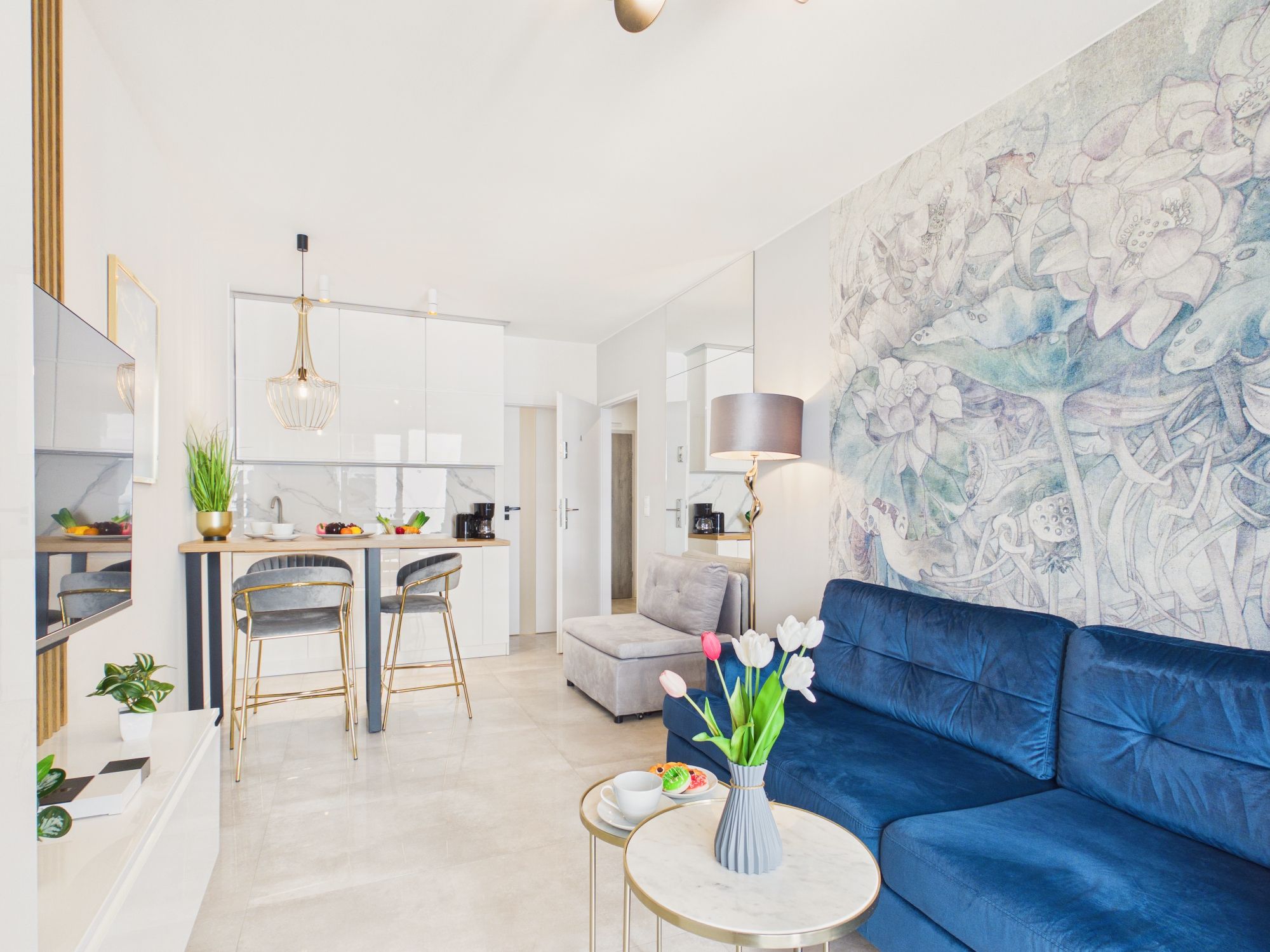 Ett modernt vardagsrum med en blå soffa, en matplats med ett kök i bakgrunden och ett stort blommotiv på väggen.