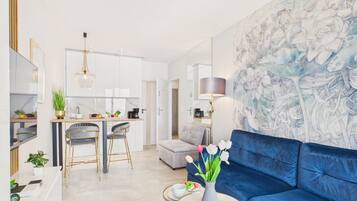 Una sala de estar moderna con un sofá azul, una zona de comedor con una cocina al fondo y una gran obra de arte floral en la pared.