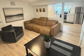 A living room with a brown leather sofa, a dark armchair, and a coffee table with a plant. A painting is on the wall, and a kitchen is visible through an open door. - Charming 4 bedroom 2 bath house in Hallandale (Hallandale)