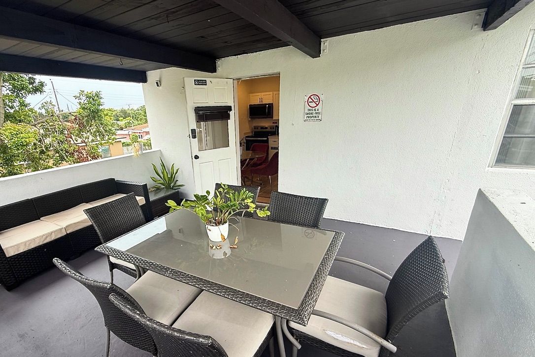 An outdoor patio with a glass-top table surrounded by wicker chairs, a potted plant, and a view of the neighborhood.