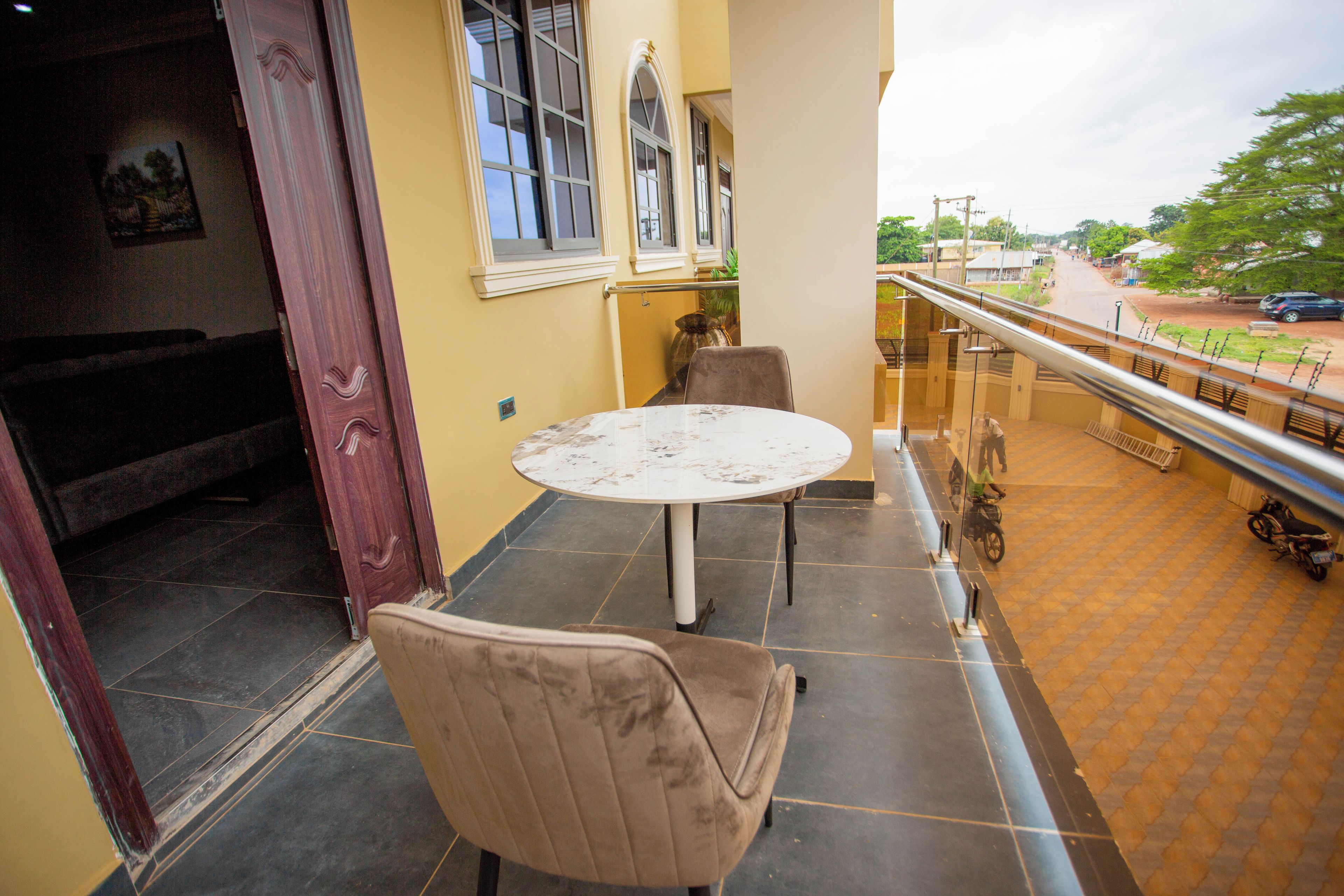 Een balkon met een ronde tafel en twee stoelen, uitkijkend op een straat met geparkeerde auto's en groen.