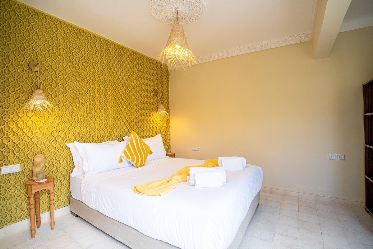 Habitació d'hotel amb paper tapet de disseny, un llit amb roba de llit blanca, una taula de nit i dues làmpades subjectes a la paret.