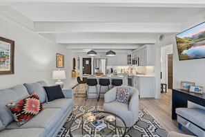 Ein modernes Wohnzimmer mit einer Couch, Sesseln, einem Couchtisch und einer Küchenzeile inklusive Kühlschrank, Mikrowelle und Barhocker.