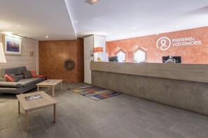Ein Empfangsbereich mit einem Holzbüro, einem grauen Sofa mit gemusterten Kissen, einem kleinen Tisch und einer Willkommensmatte.