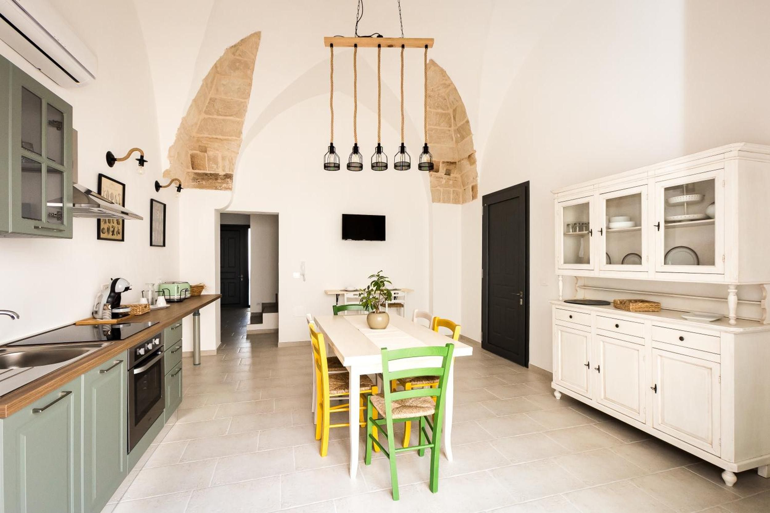 Cozinha moderna com área de refeições, destacando um arco de pedra, luminária pendente e armário com portas de vidro.