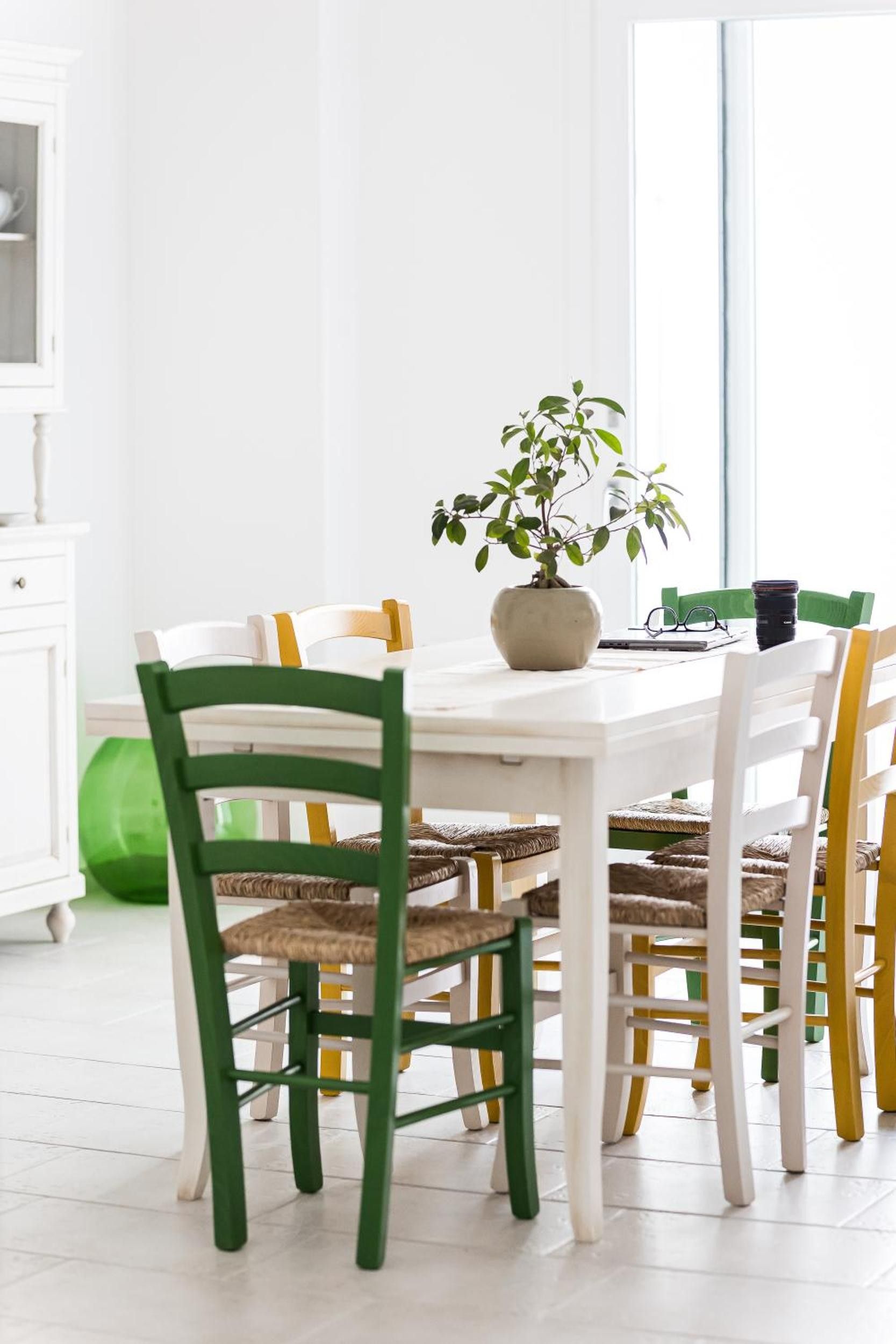 Uma sala de jantar com uma mesa branca, cadeiras verdes e amarelas, um vaso de planta e uma janela ao fundo.