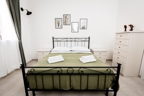 Un dormitorio ordenado con estructura de cama metálica, cubrecama verde, almohadas blancas y dos mesitas de noche con cajones blancos.