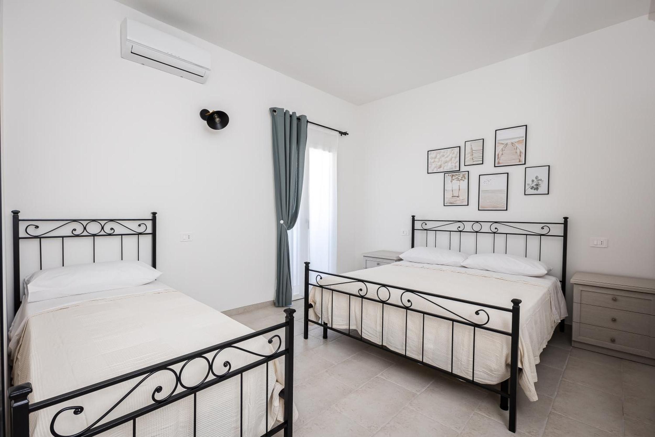 A bedroom with two beds, a window with curtains, a wall-mounted air conditioner, and several framed pictures.