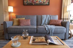 A grey sofa with multiple cushions, a glass vase, a magazine, and a lamp in a room with an orange wall and a framed artwork. - Wifi, 5m from ski slopes, view mountain, swimming pool, sauna, hammam, balcony, ski locker, 49-56m² (Flaine)